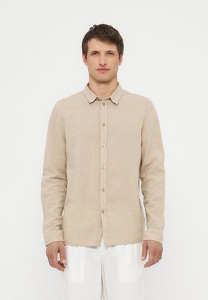 Mand iført en beige langærmet skjorte med knapper og hvide bukser, stående foran en ensfarvet hvid baggrund, vendt fremad.