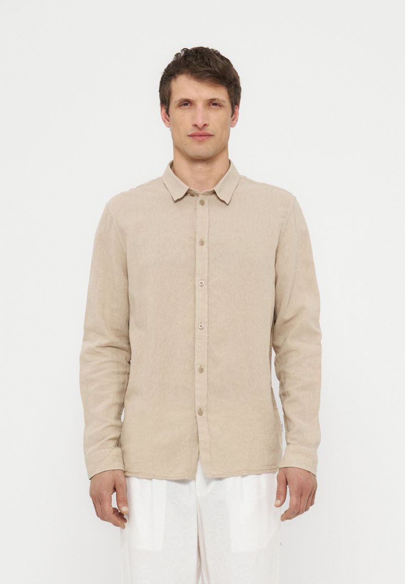 Άνδρας με μπεζ πουκάμισο μακρυμάνικο με κουμπιά και λευκό παντελόνι, στέκεται μπροστά σε απλό λευκό φόντο, στραμμένος προς τα εμπρός.