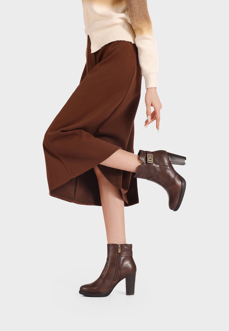 Stivali alla caviglia in pelle marrone con tacco a blocco, cerniera laterale e un dettaglio decorativo con cinturino. Indossati con una gonna midi marrone.