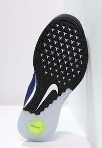 Sole tenisky Nike s černým gumovým povrchem s hexagonálními vzory, bílými akcenty a průhlednou částí, která odhaluje zelený detail.