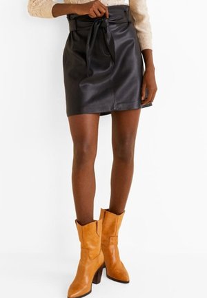 Vrouw die een zwarte leren rok met paperbag-taille draagt, vastgebonden aan de voorkant, en beige mid-kuit laarzen met hakken, staande tegen een witte achtergrond.