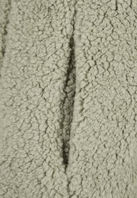 Zbliżenie na miękki, beżowy materiał sherpa fleece o gęstej, pofałdowanej strukturze i widocznym złożonym szwie na środku.