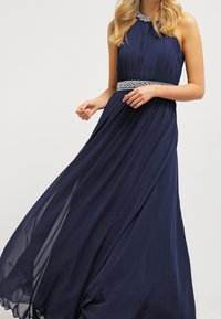 Marinblå, golvlång klänning med plisserad buste, halterneck och dekorerad midja. Flödande, lätt tyg förbättrar rörelse.