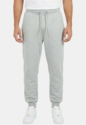 Homme portant un pantalon de survêtement gris clair à cordon et des baskets blanches, debout devant un fond blanc uni.