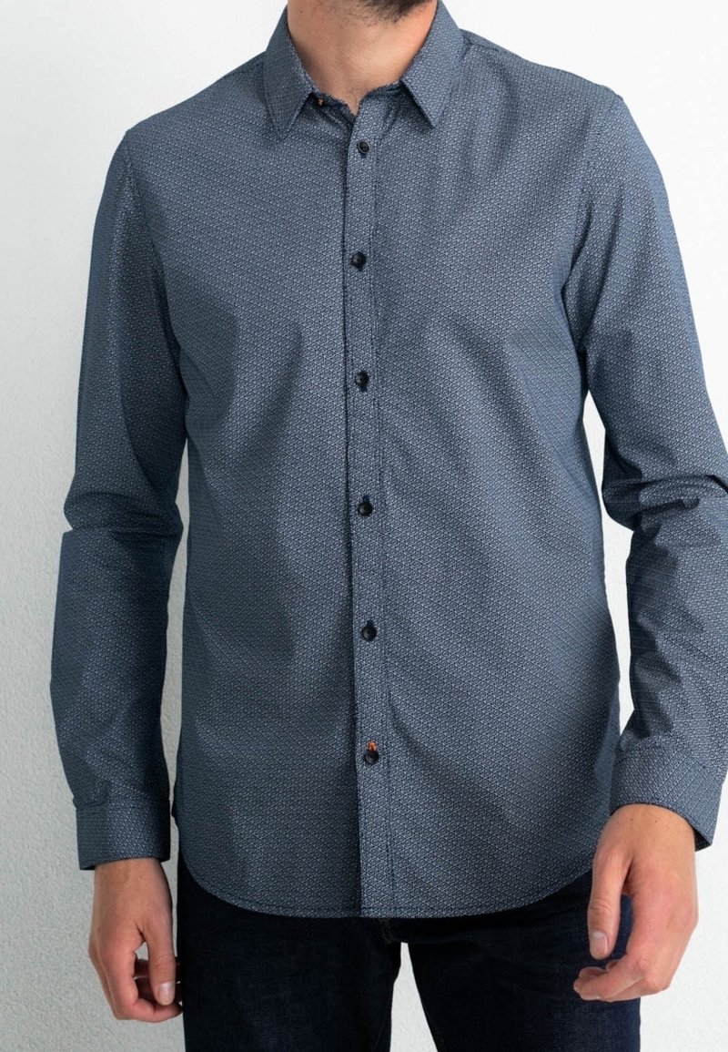 Homme portant une chemise cintrée bleue à manches longues à motifs avec un pantalon noir sur un fond uni.