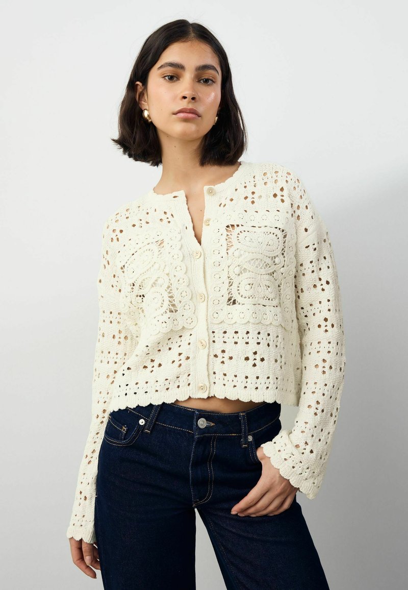 Cardigan en tricot ouvert crème avec un ourlet en forme de vagues, fermeture à boutons et motifs en dentelle complexes. Porté sur un jean en denim foncé.