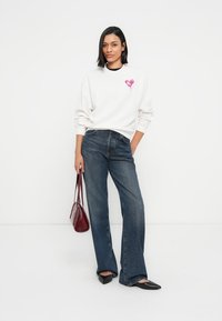 Λευκό φούτερ με ροζ καρδιά, συνδυασμένο με σκούρο μπλε τζιν φαρδύ. Το μοντέλο κρατάει μπορντό τσάντα και φοράει μαύρα μπαλαρίνες.