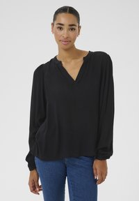 Black long-sleeve blouse with a V-neck, pleated shoulders, textured fabric, and elastic cuffs. Worn with blue jeans.