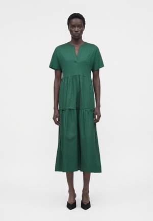 Femme portant une robe midi verte à manches courtes, col en V et jupe à volants, associée à des talons noirs pointus, debout sur un fond blanc.