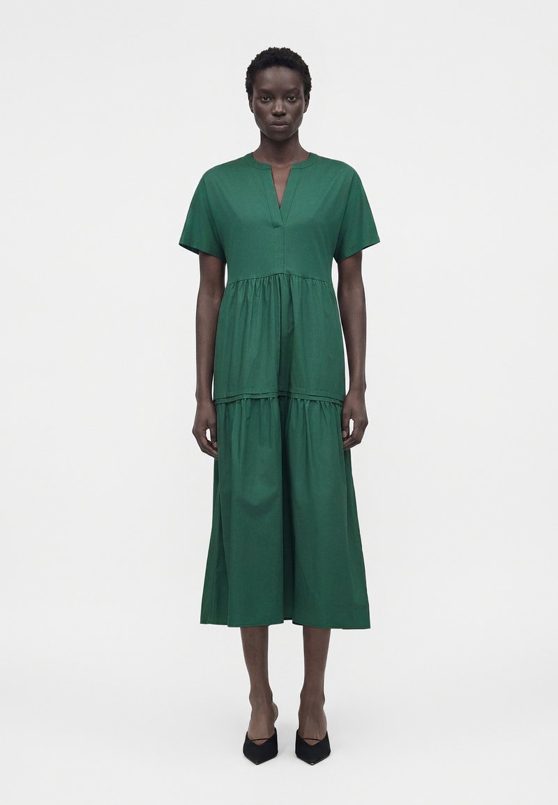 Γυναίκα με πράσινο midi φόρεμα με κοντά μανίκια, V-λαιμόκοψη και στρωτά μανίκια, συνδυασμένο με μαύρες μυτερές γόβες, στέκεται σε λευκό φόντο.