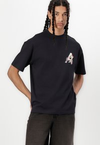 Sort bomulds T-shirt med rund halsudskæring, korte ærmer og et blomsterprydet bogstav "A" design på venstre bryst. Glat tekstur.