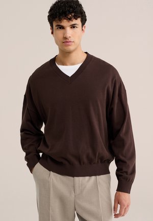 Joven con cabello rizado lleva un suéter de cuello en V marrón oscuro sobre una camisa blanca y pantalones beige, de pie con una mano en el bolsillo.