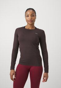 Brūns sporta krekls ar garām piedurknēm un apaļu izgriezumu, izgatavots no gludas auduma. Uz krūšu ir mazs logotips. Apvienots ar sarkaniem legingiem.