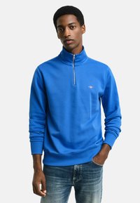 Felpa blu con zip e colletto alto, con polsini a coste. Presenta un ricamo del logo sul petto. Abbinata a jeans blu strappati.