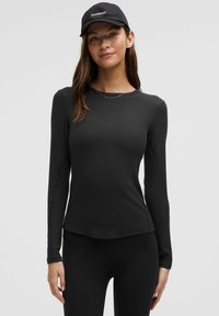 Zwarte lange mouw top van gladde stof, met een ronde hals en naaddetails, gecombineerd met zwarte leggings en een pet.