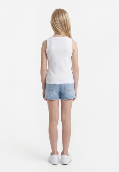 Kind met lang blond haar, draagt een wit mouwloos shirt, lichtblauwe denim shorts en witte sneakers, staand met de rug naar voren.