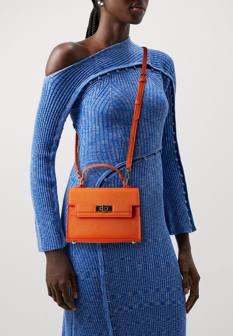 Femme portant une robe bleue à épaules dénudées et côtelée, et portant un petit sac à main orange vif avec une poignée supérieure et une bandoulière.