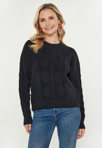 Femeie zâmbitoare cu păr blond, purtând cercei mari în formă de floare roșie, un pulover texturat negru și blugi, pe un fundal simplu.