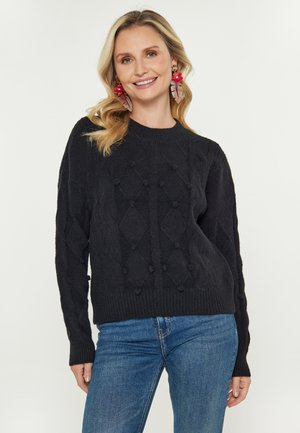 Femeie zâmbitoare cu păr blond, purtând cercei mari în formă de floare roșie, un pulover texturat negru și blugi, pe un fundal simplu.