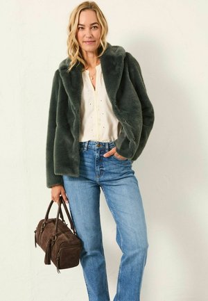 Faux fur, dark green jacket with a shawl collar, worn over a cream blouse, blue jeans, and holding a brown suede handbag.