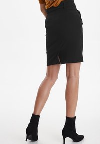 Jupe crayon noire avec fente latérale, présentant une texture lisse et un design ajusté. Associée à des bottines noires, présentée de dos.