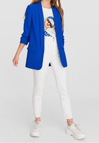 Kungsblå blazer, vit grafisk t-shirt, vita slim-fit jeans och vita sneakers. Blazern har slagna kragar och uppvikta ärmar.