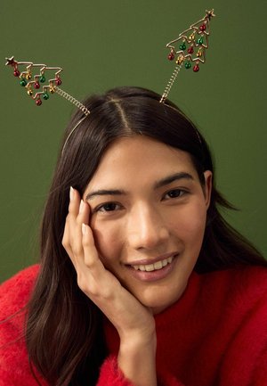 Young woman in red sweater smiling, resting her face on her hand, wearing a headband with festive Christmas tree decorations.