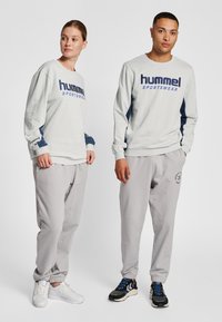 Jasnoszara bluza z ciemnoniebieskimi akcentami i nadrukiem "hummel SPORTSWEAR"; w zestawie z dopasowanymi szarymi dresami oraz białymi i czarnymi sneakersami.