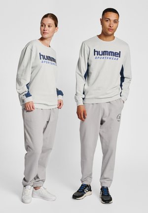 Ljusgrå sweatshirt med mörkblå detaljer och tryck med texten "hummel SPORTSWEAR"; matchad med grå mjukisbyxor och vita och svarta sneakers.