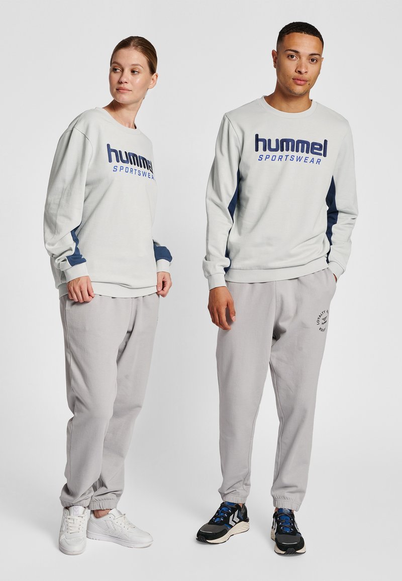 Jasnoszara bluza z ciemnoniebieskimi akcentami i nadrukiem "hummel SPORTSWEAR"; w zestawie z dopasowanymi szarymi dresami oraz białymi i czarnymi sneakersami.