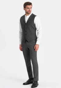 Grijze wollen pak met een aansluitende vest, vijf knopen en notch revers. Gepaarde met grijze broek en zwarte leren schoenen. Wit overhemd eronder.