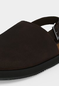 Σανδάλι από σκούρο καφέ σουέντ με κλειστή μύτη, μαύρη σόλα και ρυθμιζόμενο ασημένιο λουράκι με αγκράφα στο τακούνι.