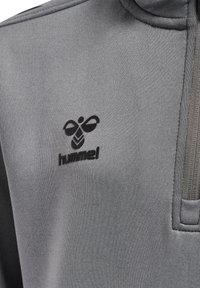 Felpa grigia con colletto alto e zip, caratterizzata da un logo di ape nera e il testo "hummel", realizzata in un tessuto liscio con una leggera trama.