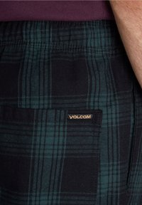 Shorts à carreaux en tissu vert foncé et noir, avec une poche latérale ornée d'une étiquette au logo Volcom. Taille élastique pour plus de confort.