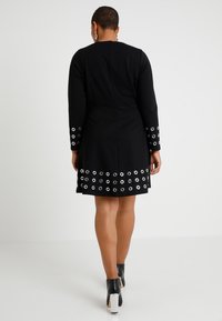 Lyhythiuksinen nainen, joka wear musta pitkähihaista mekkoa, jossa on hopeiset rei'itetyt yksityiskohdat hihoissa ja helmassa, kävelee poispäin valkoista taustaa vasten.