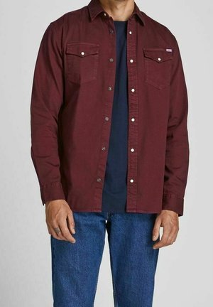 Homme portant une chemise bordeaux à boutons par-dessus un t-shirt bleu marine et un jean bleu, debout devant un fond uni.