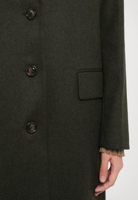 Svetle olivově zelený vlněný kabát s šesti knoflíky, náprsní kapsou a texturovaným manžetou z podkladového oděvu.