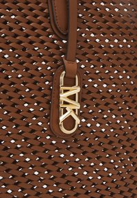 Bruine geweven handtas met een zachte leren schouderriem en een gouden MK-logo accent. Gedetailleerde textuur voegt een driedimensionale uitstraling toe.