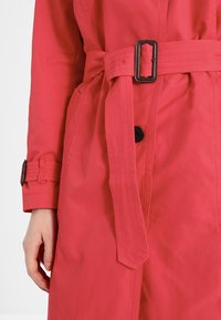 Trench-coat rouge avec ceinture à la taille dotée d'une boucle noire rectangulaire, fermeture par boutons et sangle de manche réglable avec boucle assortie.