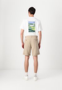 Fehér póló színes bélyeg grafikával a hátsó oldalon, bézs rövidnadrággal párosítva. A rövidnadrágok felgörgött szegéllyel és két hátsó zsebbel rendelkeznek.