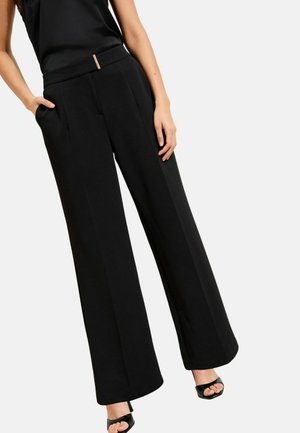 Žena oblečená v čiernych širokých nohaviciach s bočnými vreckami a čiernom bezrukávovom topu, stojaca s jednou rukou vo vrecku a na nohách má čierne sandále s otvorenou špičkou.