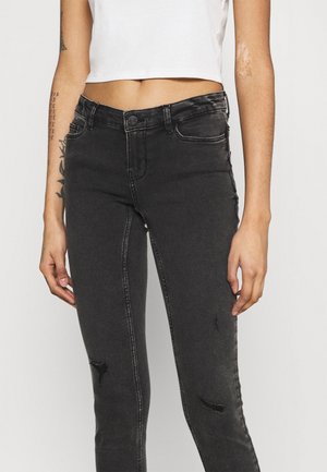 Close-up af en person iført sorte distressed skinny jeans og en hvid cropped ærmeløs top mod en ensfarvet baggrund.