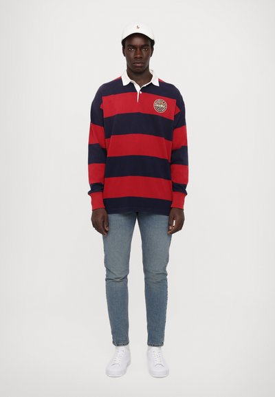 Tricou de rugby în dungi bleumarin și roșii, cu guler alb, având un badge pe piept; asociat cu blugi albaștri deschis și adidași albi.