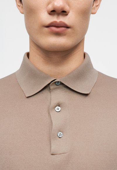 Μπεζ πόλο πουκάμισο με υφασμάτινη υφή, διαθέτει απαλό γιακά και κλείσιμο με τρία κουμπιά σε αντίστοιχους χρωματισμούς. Μινιμαλιστικός σχεδιασμός χωρίς σχέδια.