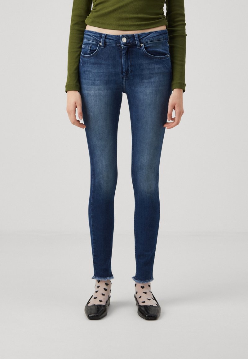 Jeans skinny bleu marine avec une taille mi-haute, présentant des poches avant et un ourlet légèrement effiloché. Portés avec des chaussettes à motifs et des ballerines.