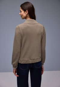 Mujer con cabello largo y castaño, que lleva pendientes de aro dorados, una chaqueta beige con cuello y vaqueros azul oscuro, de espaldas contra un fondo gris.