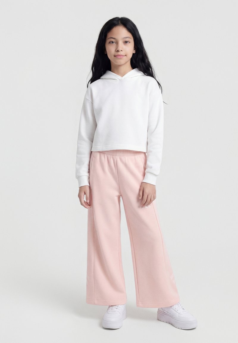 Fille debout portant un sweat à capuche blanc court, un pantalon large rose clair et des baskets blanches sur un fond clair uni.