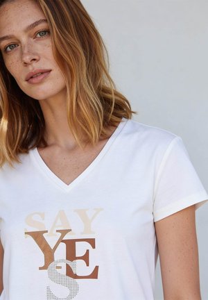 Woman with shoulder-length light brown hair wearing a white V-neck T-shirt with the text "SAY YES" in beige and brown letters.