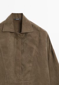 Giacca in suede marrone con un ampio colletto e una profonda patta frontale a V. Texture liscia con dettagli di cucitura visibili, design minimalista.