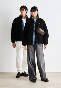 Two adults wearing black fleece jackets over blue plaid shirts, one with white pants and sunglasses, the other with gray jeans and a brown crossbody bag.